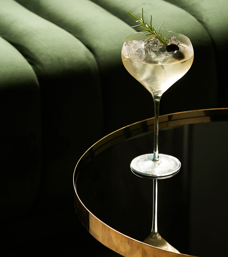 close up of coupe cocktail glass on a copper rimmed table with a green sofa in background