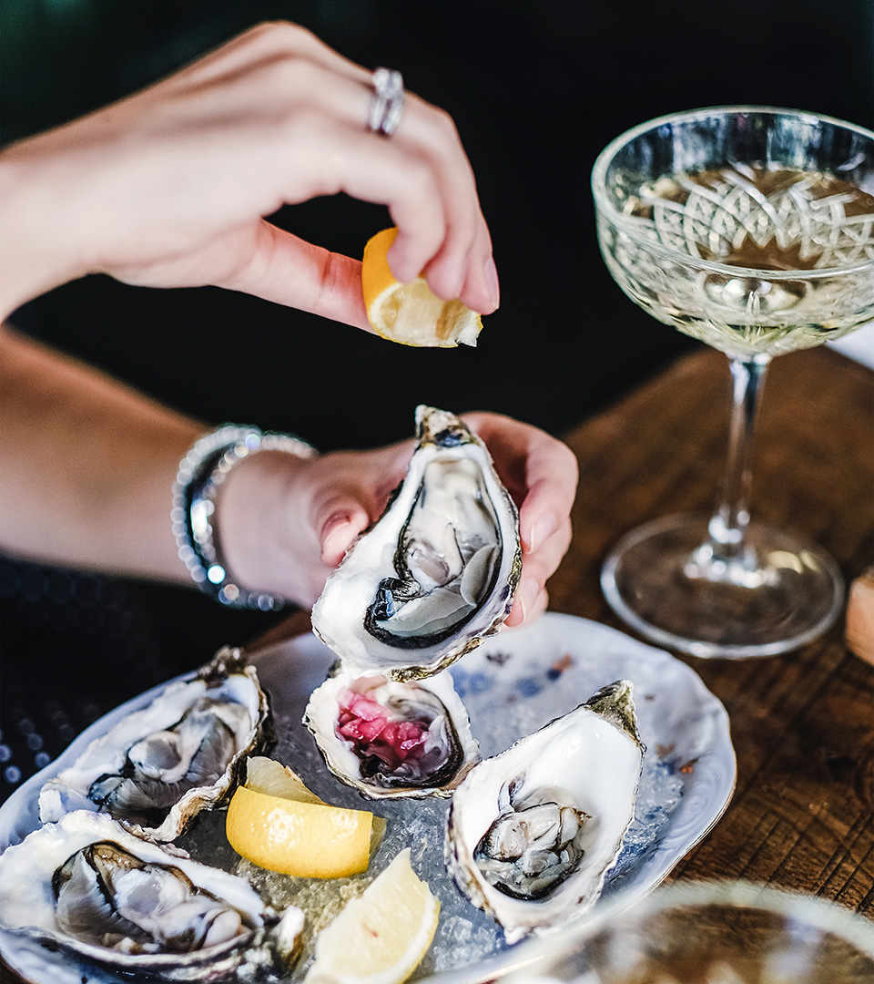 Hands of woman squeezing lemon juice to Irish oysters in plate with ice over glass of champagne by best boston ad agency